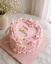 Heart-shaped pink birthday cake with satin ribbons, golden accents and detailed frosting