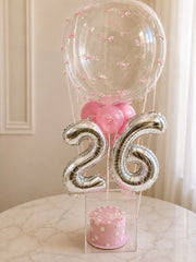 Petal Grace pink birthday cake with floral details, silver number balloons, pink balloons, and clear acrylic display