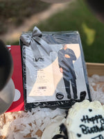 Load image into Gallery viewer, A packaged dark navy or black long-sleeve shirt set with a printed model photo and a gray ribbon bow, placed in a gift box with shredded paper. A blurred outdoor background and part of a birthday cake are visible around it.