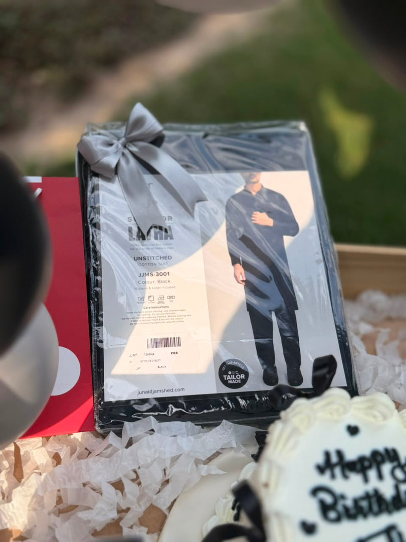 A packaged dark navy or black long-sleeve shirt set with a printed model photo and a gray ribbon bow, placed in a gift box with shredded paper. A blurred outdoor background and part of a birthday cake are visible around it.