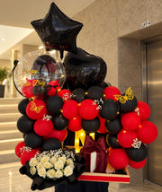 Red and black birthday balloon arrangement with black star and heart balloons, ivory roses, fairy lights and a gift box with a burgundy bow.

