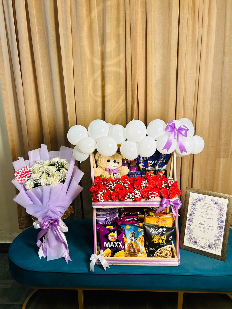 Wedding snack hamper with red roses, a teddy bear and white balloons beside a lavender-wrapped ivory rose bouquet and framed wedding keepsake.