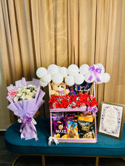 Wedding snack hamper with red roses, a teddy bear and white balloons beside a lavender-wrapped ivory rose bouquet and framed wedding keepsake.