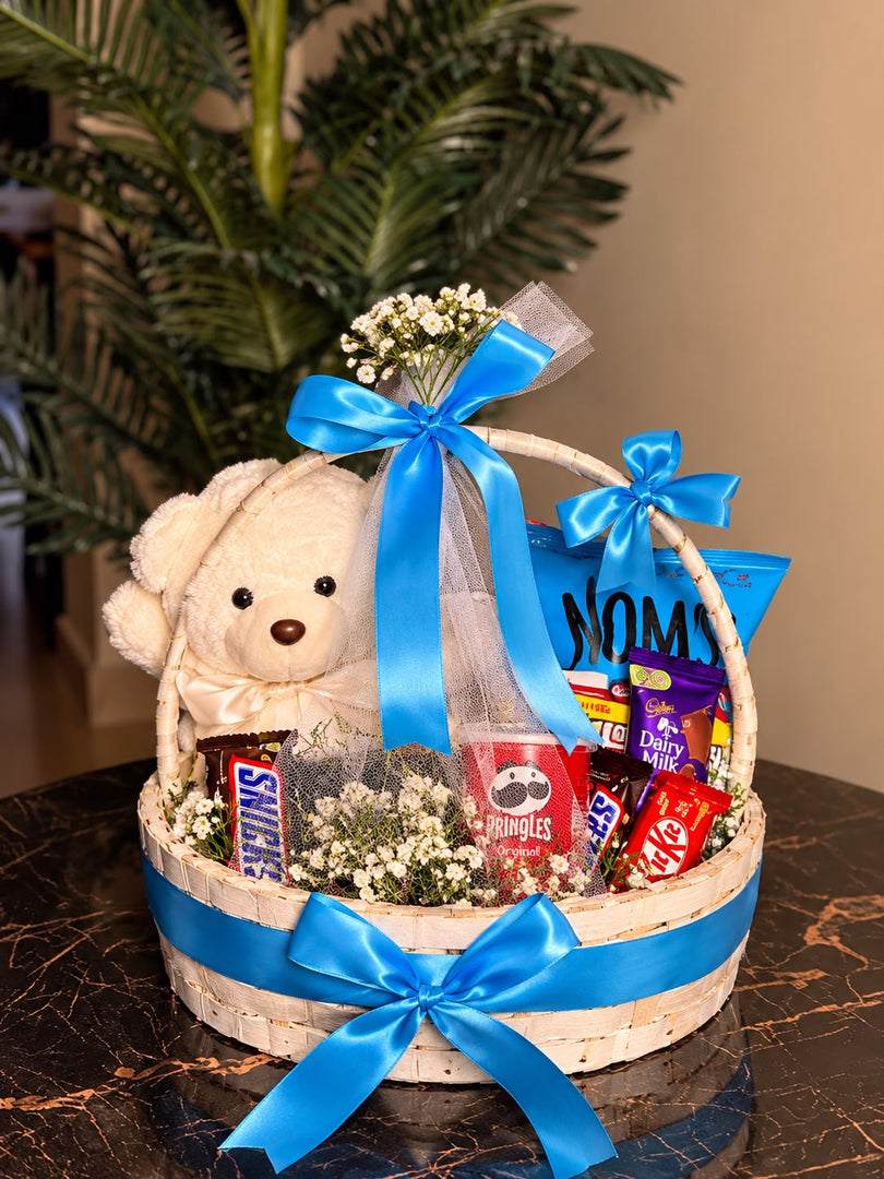 Cream teddy bear and assorted chocolates in a woven gift basket decorated with blue satin ribbons and white flowers.
