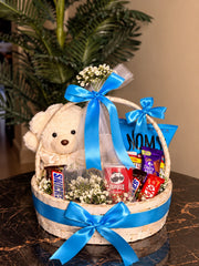 Cream teddy bear and assorted chocolates in a woven gift basket decorated with blue satin ribbons and white flowers.