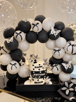 Load image into Gallery viewer, Three-tier black and white birthday cake with hearts and butterflies beneath a matching balloon arch with black bows and clear confetti balloons.