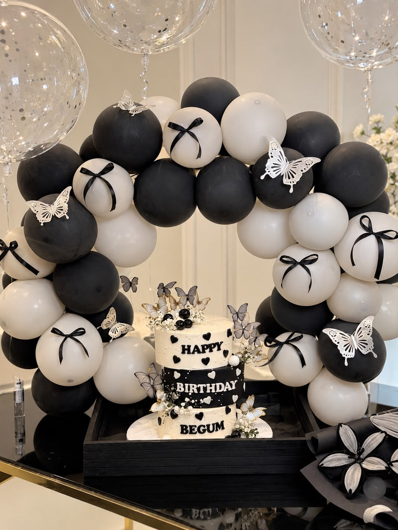 Three-tier black and white birthday cake with hearts and butterflies beneath a matching balloon arch with black bows and clear confetti balloons.