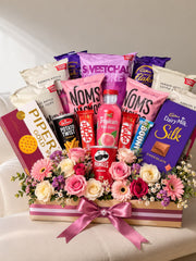 Gift hamper filled with assorted snacks, chocolate bars, juice, pink and white roses, and a pink satin bow.