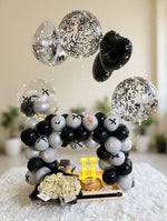Load image into Gallery viewer, Black and silver birthday balloon arrangement with cream roses, a small cake, fairy lights, and an illuminated letter H in a wooden tray.