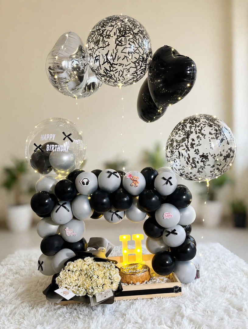 Black and silver birthday balloon arrangement with cream roses, a small cake, fairy lights, and an illuminated letter H in a wooden tray.