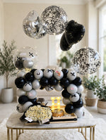 Load image into Gallery viewer, Cream rose bouquet in dark wrapping beside a birthday cake and glowing letter H beneath a black and silver balloon arch.
