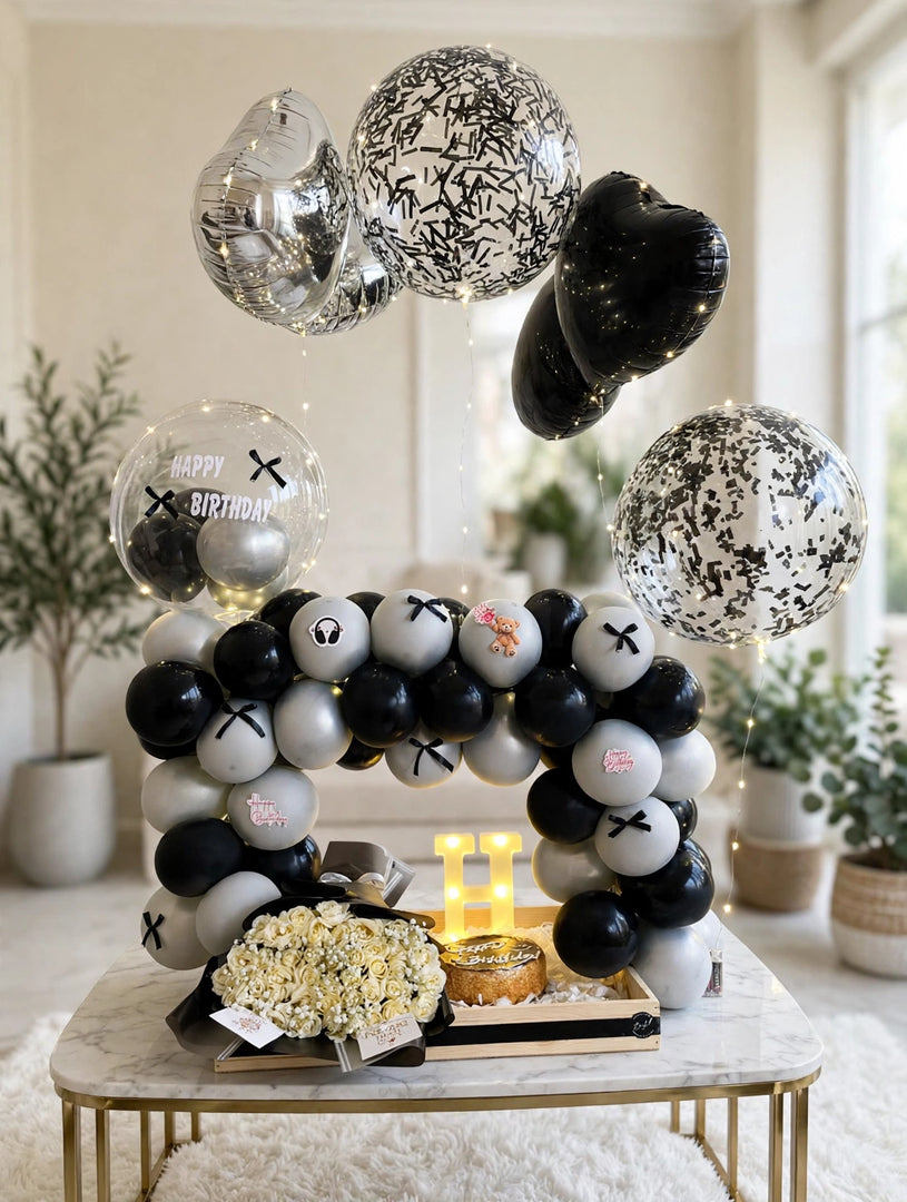 Cream rose bouquet in dark wrapping beside a birthday cake and glowing letter H beneath a black and silver balloon arch.