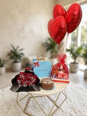 Birthday gift Pack with red roses in black wrapping, a round cake, a snack hamper, gift bags, and three red heart balloons.