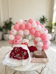 Birthday gift pack with a pink and white balloon arch, red rose bouquet, and small cake arranged in a pink-edged tray.